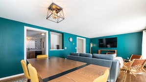 Un salón moderno con paredes verde azulado, una mesa de comedor de madera, sillas amarillas, un sofá gris y un televisor fijado a la pared.