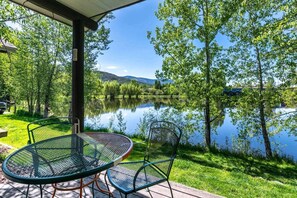 An outdoor seating area with a table and chairs overlooking a calm lake, surrounded by trees and a clear sky.