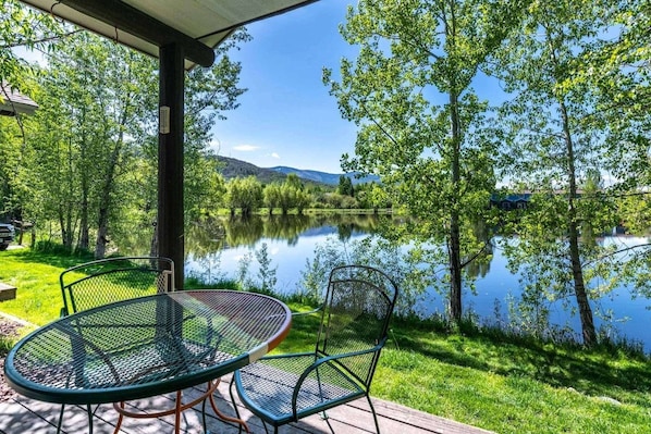 An outdoor seating area with a table and chairs overlooking a calm lake, surrounded by trees and a clear sky.