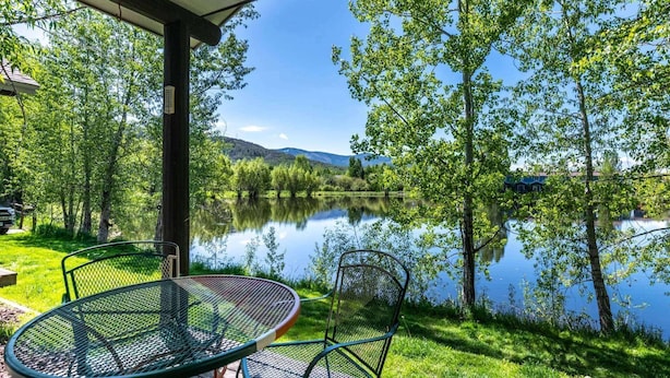 Un patio junto al lago con dos mesas y sillas de metal, rodeado de exuberante vegetación y un lago tranquilo que refleja el cielo.