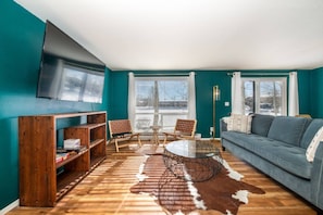 A living room with teal walls, a blue sofa, a wooden bookshelf, a glass coffee table, and a cowhide rug.