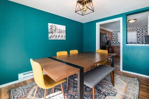 Une pièce aux murs de couleur turquoise, avec une table à manger en bois, des chaises jaunes et un banc. On y trouve un tapis à motifs sur le sol, une décoration murale en noir et blanc, et une cuisine visible à travers une porte ouverte.