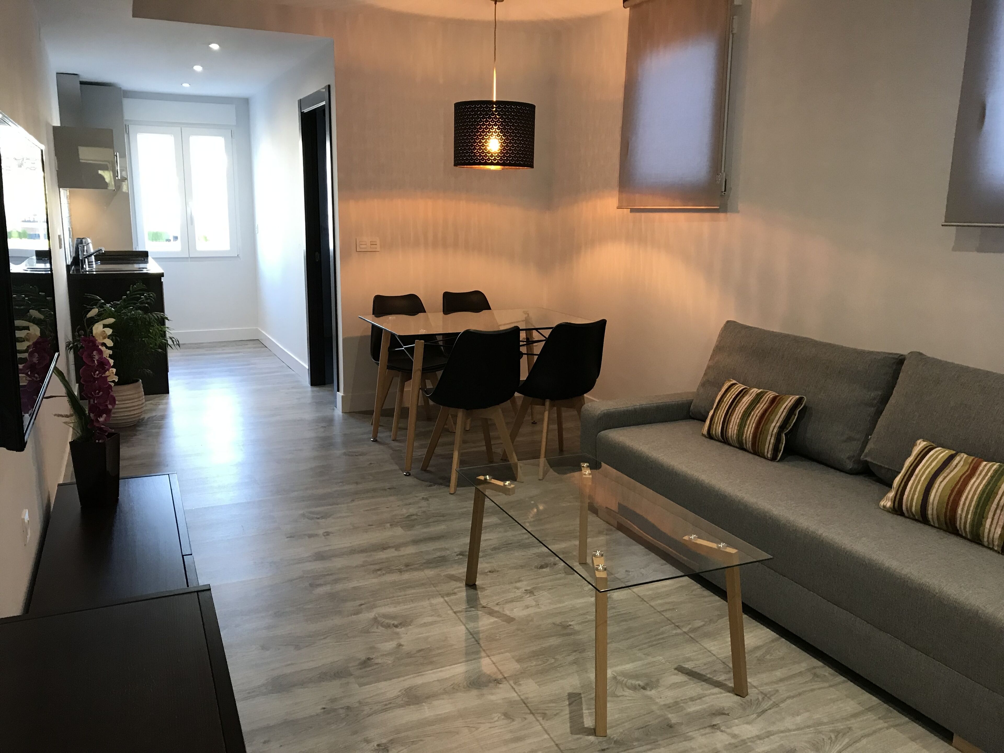 Ein modernes Wohnzimmer mit Glastisch, grauem Sofa und schwarzen Stühlen. Ein Essbereich mit Tisch und Stühlen ist ebenfalls vorhanden, und im Hintergrund ist eine Küche zu sehen.