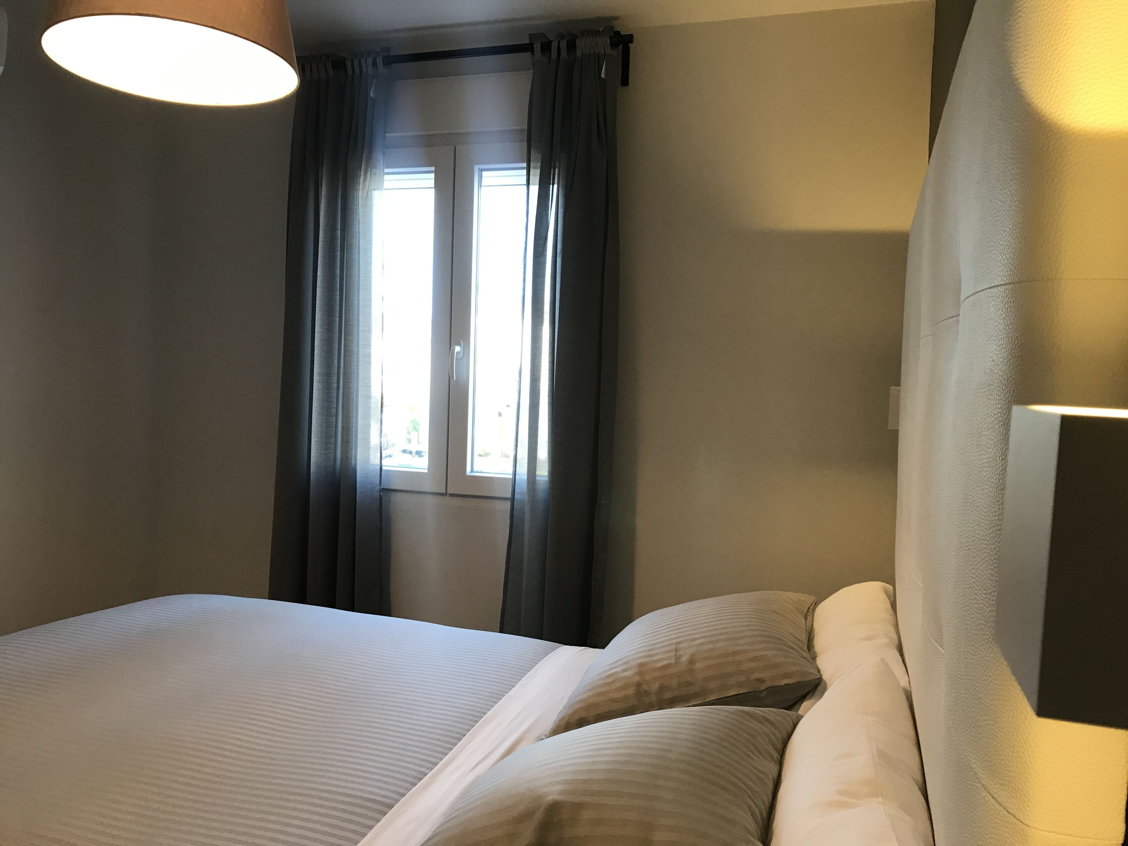 A hotel room with a bed, curtains, a window, and a wall-mounted lamp.