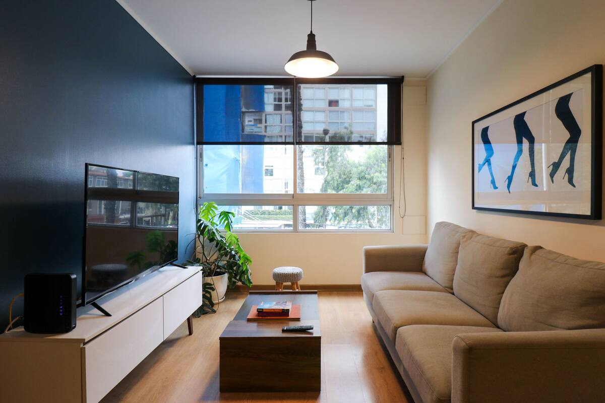 Ein modernes Wohnzimmer mit großem Fernseher, einer Couch, einem Couchtisch und einem Fenster mit Blick auf Gebäude und Bäume.