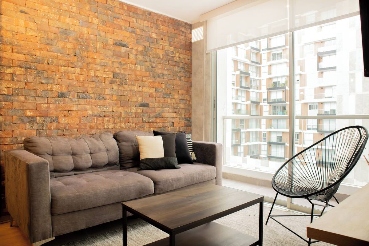Ein modernes Wohnzimmer mit Backsteinwand, einem grauen Ecksofa, einem schwarzen Sessel und einem großen Fenster mit Blick auf ein Wohngebäude.