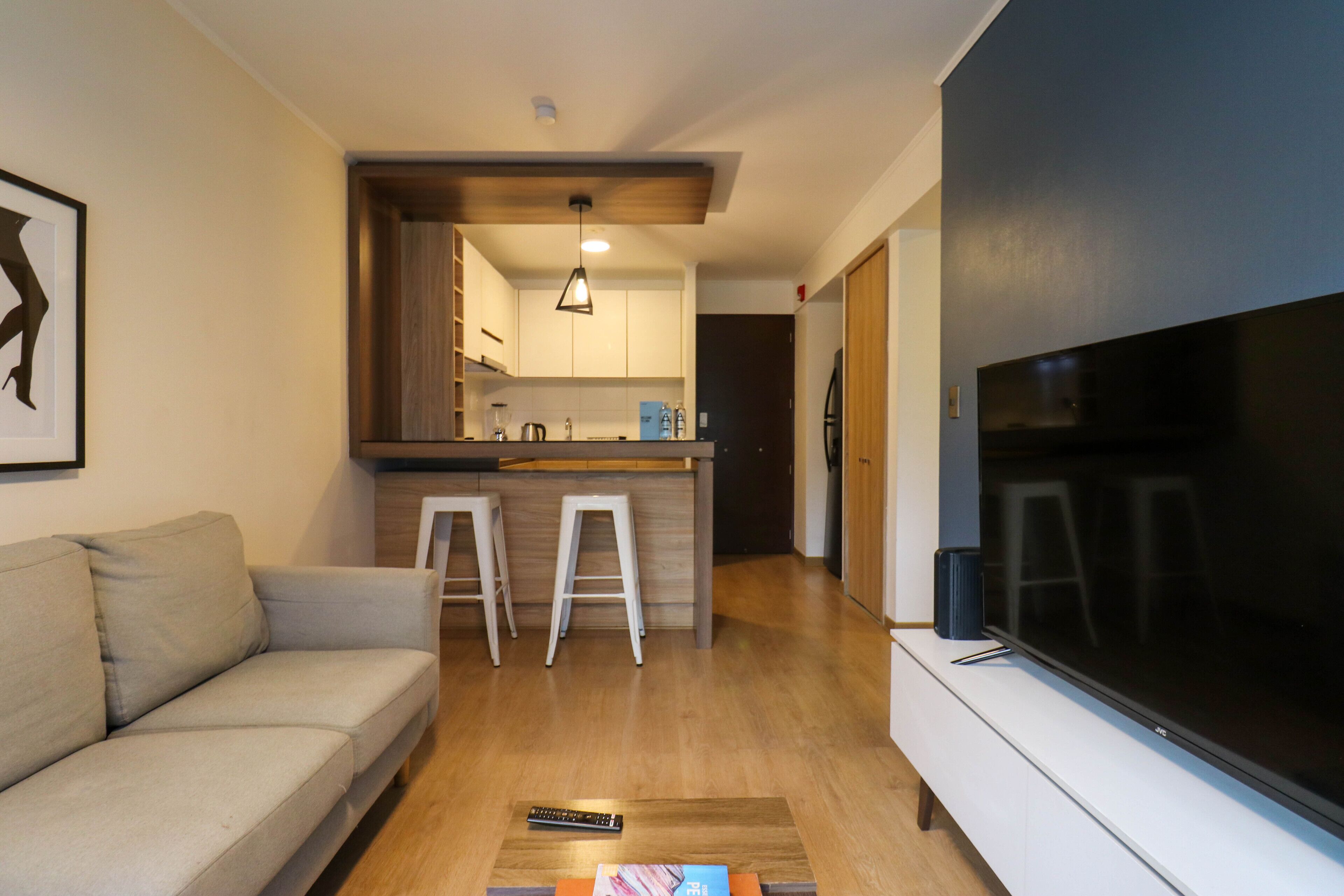 En moderne stue med sofa, sofabord med fjernbetjeninger, fjernsyn og et køkkenområde med barstole.