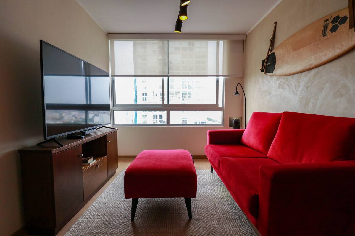 Ein Wohnzimmer mit einem großen Flachbildfernseher, einem roten Sofa, einem roten Hocker und einem Surfbrett an der Wand.