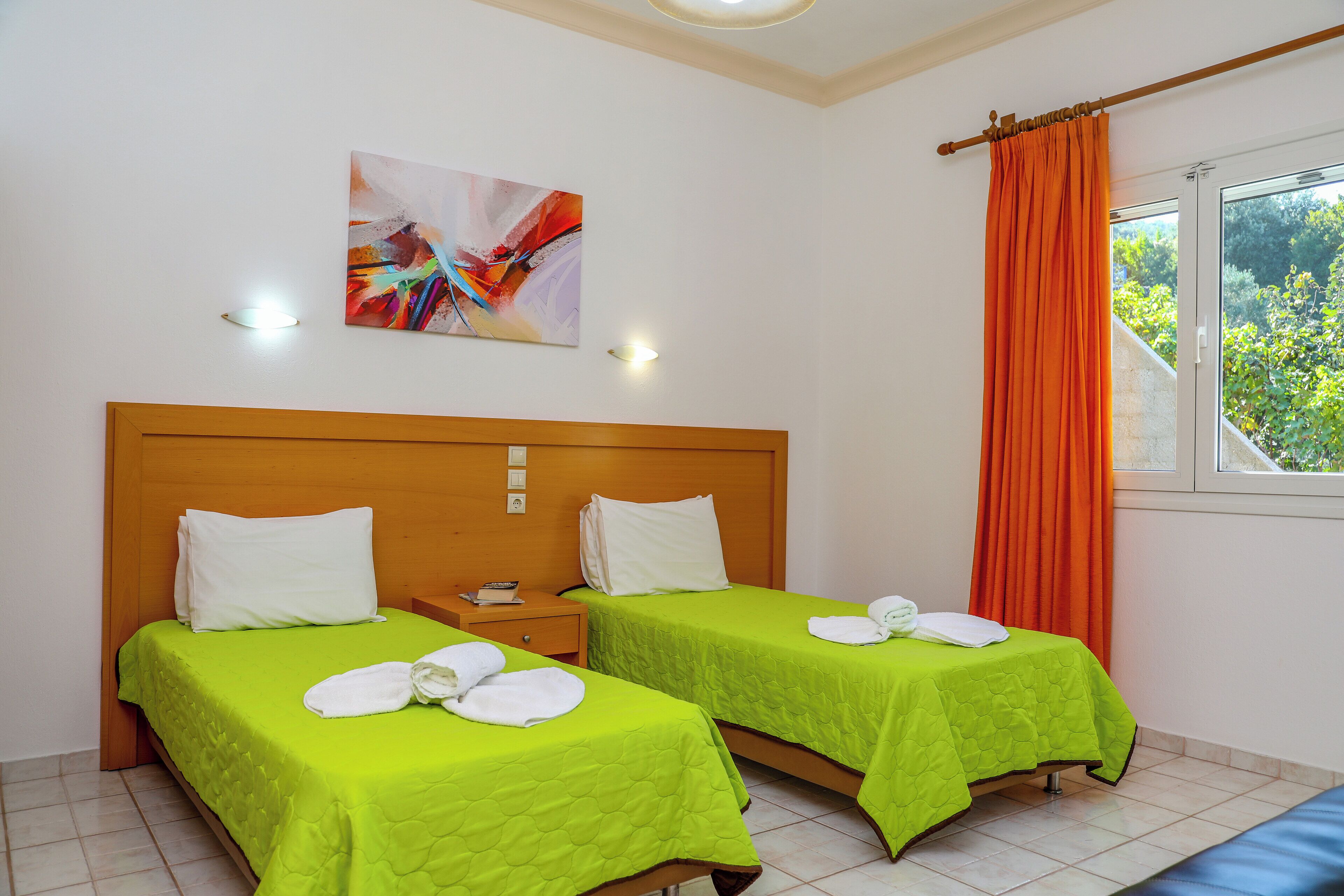 A hotel room with two beds, each with a green bedspread and white pillows, a wooden headboard, a painting on the wall, and a window with orange curtains.