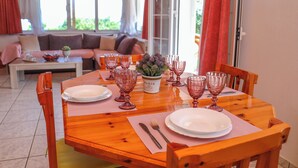 Una mesa de comedor con platos, cubiertos y vasos de vidrio rosa, rodeada de sillas de madera en una habitación luminosa, con un sofá y una ventana.