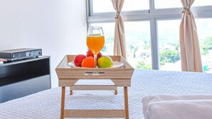 Quarto de hotel com cama, mesa de cabeceira com bandeja de frutas e um copo de suco de laranja, e uma janela com cortinas.