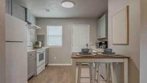 A compact kitchen with white cabinets, a central island, and a window with blinds. - 2-Bedroom Cottage w/ KING bed (Edmond)