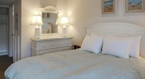 Una cama bien hecha con cabecera blanca y dos almohadas, una mesita de noche con lámpara, y un espejo encima.
