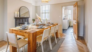 Comedor con mesa de madera dispuesta para seis comensales, sillas blancas, candelabro, chimenea y vista a la cocina.