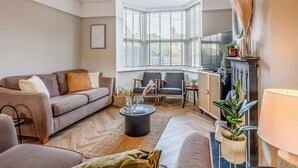 Una sala de estar con un sofá, sillones, una mesa de centro y un televisor. Hay una ventana con persianas, una chimenea y una lámpara.