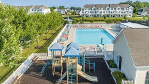 Une aire de jeux avec un toboggan et des structures d’escalade, une piscine et une clôture blanche qui entoure l’ensemble.