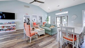 Un vaste salon doté d’un ventilateur de plafond, d’un téléviseur, d’une bibliothèque, d’un coin repas avec une table et des chaises, et d’une cuisine visible à travers une porte ouverte.