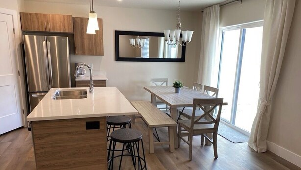 A modern kitchen with a central island, stainless steel appliances, wooden cabinets, and a dining area with a table and chairs. - Gorgeous New Townhome with Hot Tub (Kanab)
