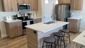 A modern kitchen with wooden cabinets, stainless steel appliances, a central island with stools, and pendant lighting. - Gorgeous New Townhome with Hot Tub (Kanab)