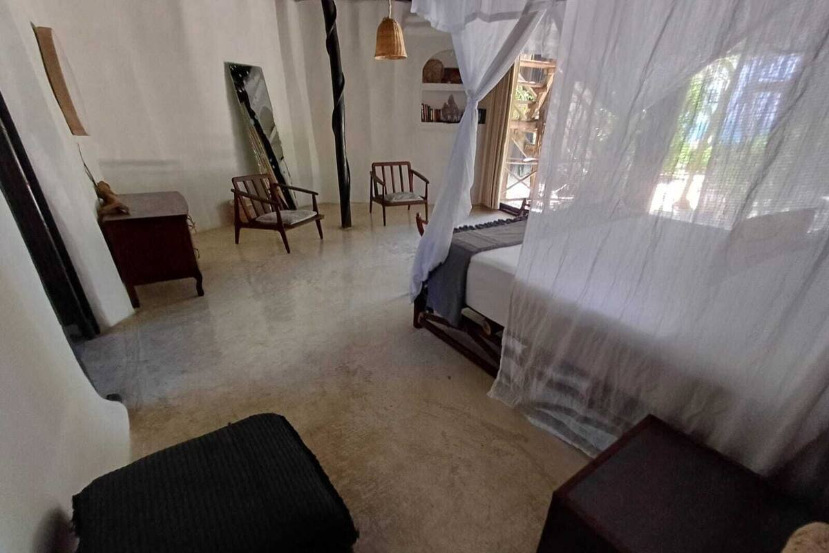 A room with a bed covered by a sheer white curtain, a wooden chair, a small table, and a large mirror on the wall.