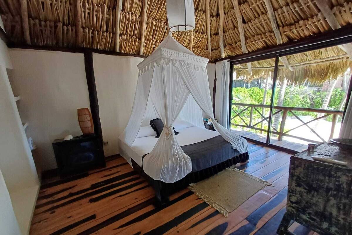A room with a bed under a white canopy, wooden flooring, and a large window with a view of greenery.