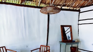 Habitació amb una taula rodona de fusta i quatre cadires, un llum penjant teixit, una taula petita amb un mirall i llibres, i un finestral gran amb vista a arbres.