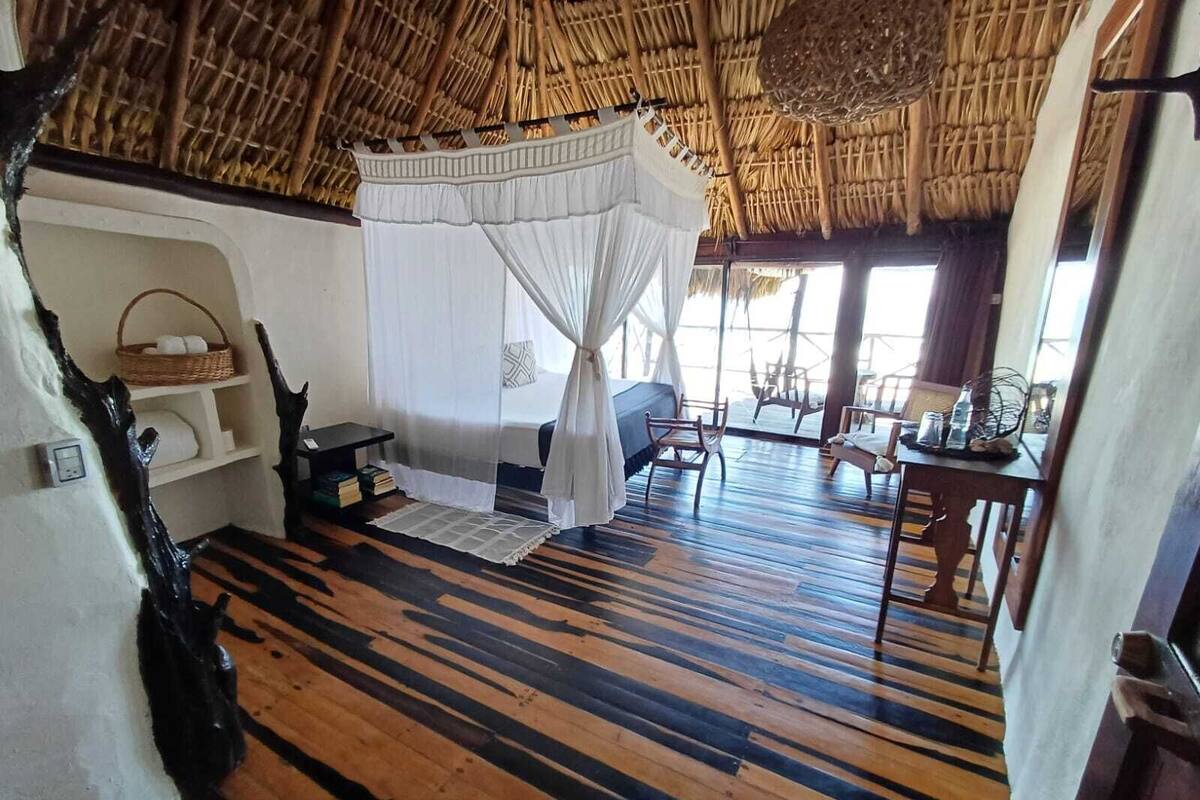 A room with a thatched roof, a bed with a canopy, wooden flooring, and a view of the sea.