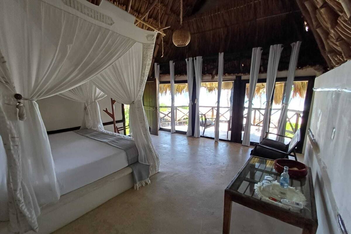 A room with a bed under a canopy, a glass table with a bottle, and a view of the outdoors.