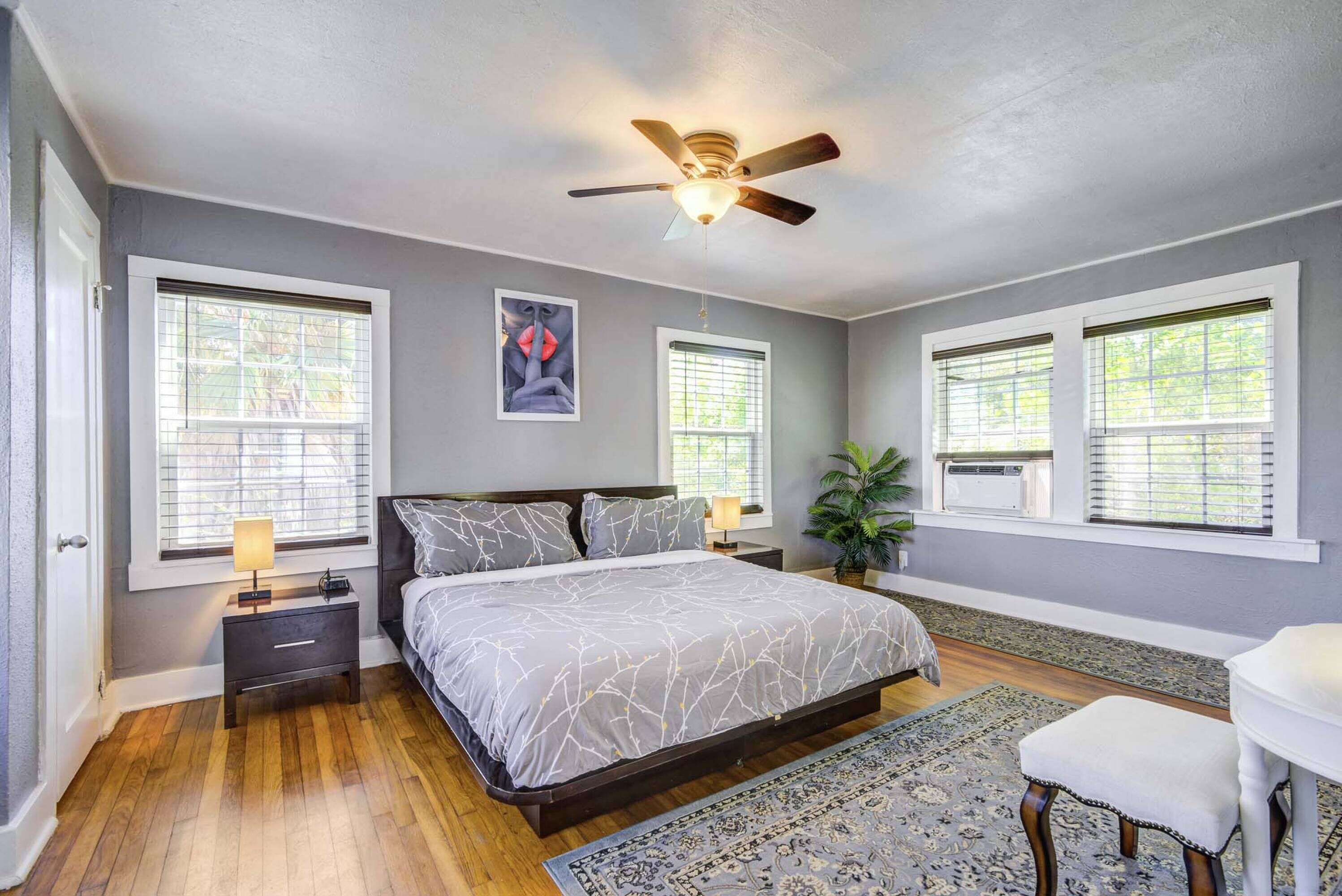 A bedroom with a bed, nightstands, a ceiling fan, and large windows.