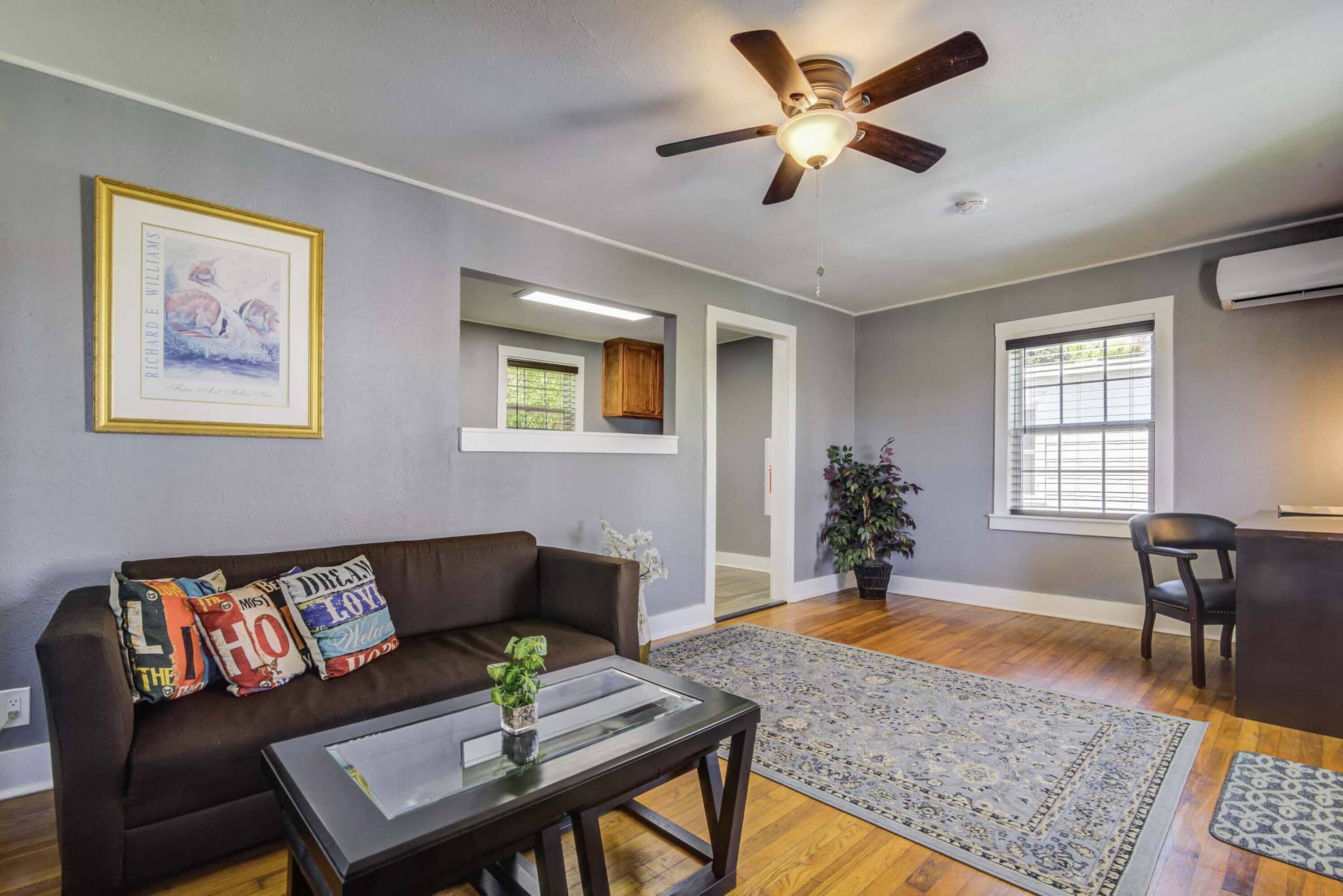 A living room with a brown sofa, a glass coffee table, a ceiling fan, and a large window with blinds.