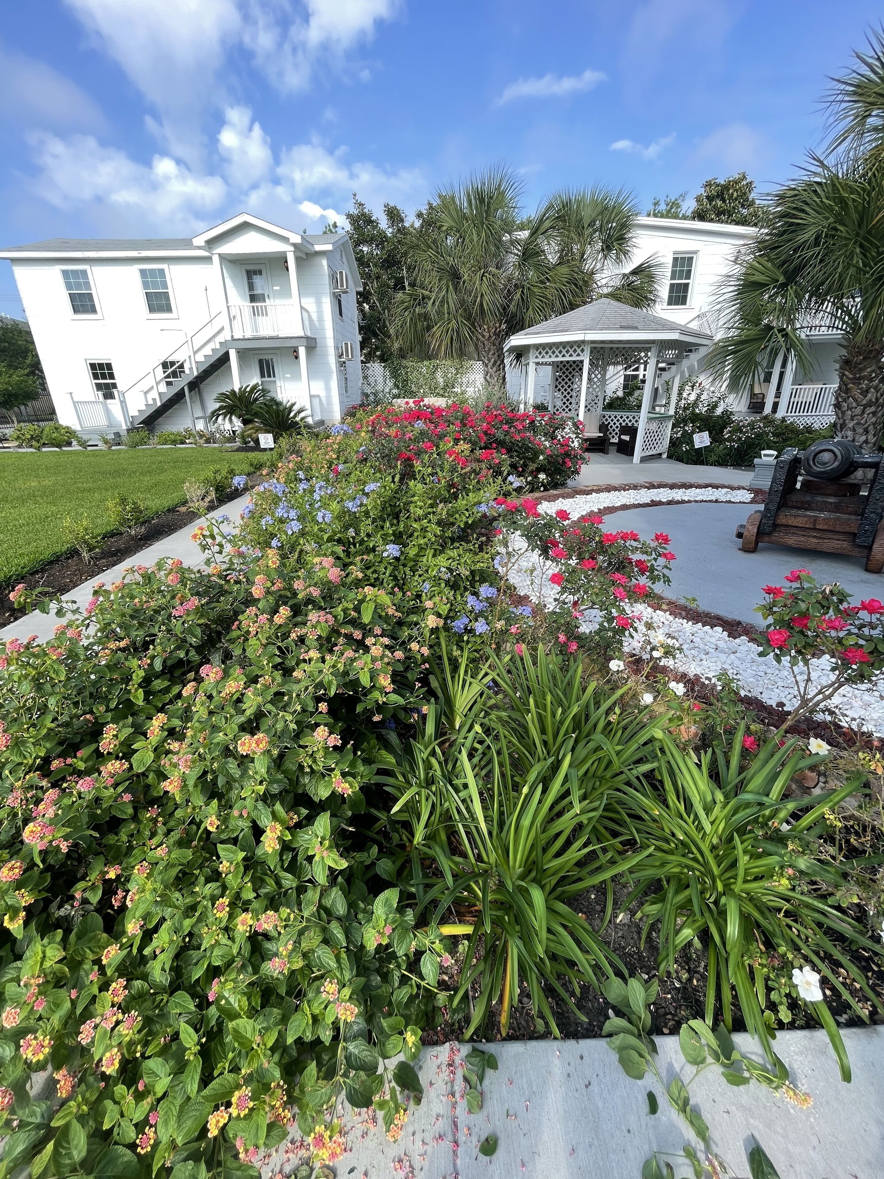A white building with a covered porch and a garden with various plants and flowers.