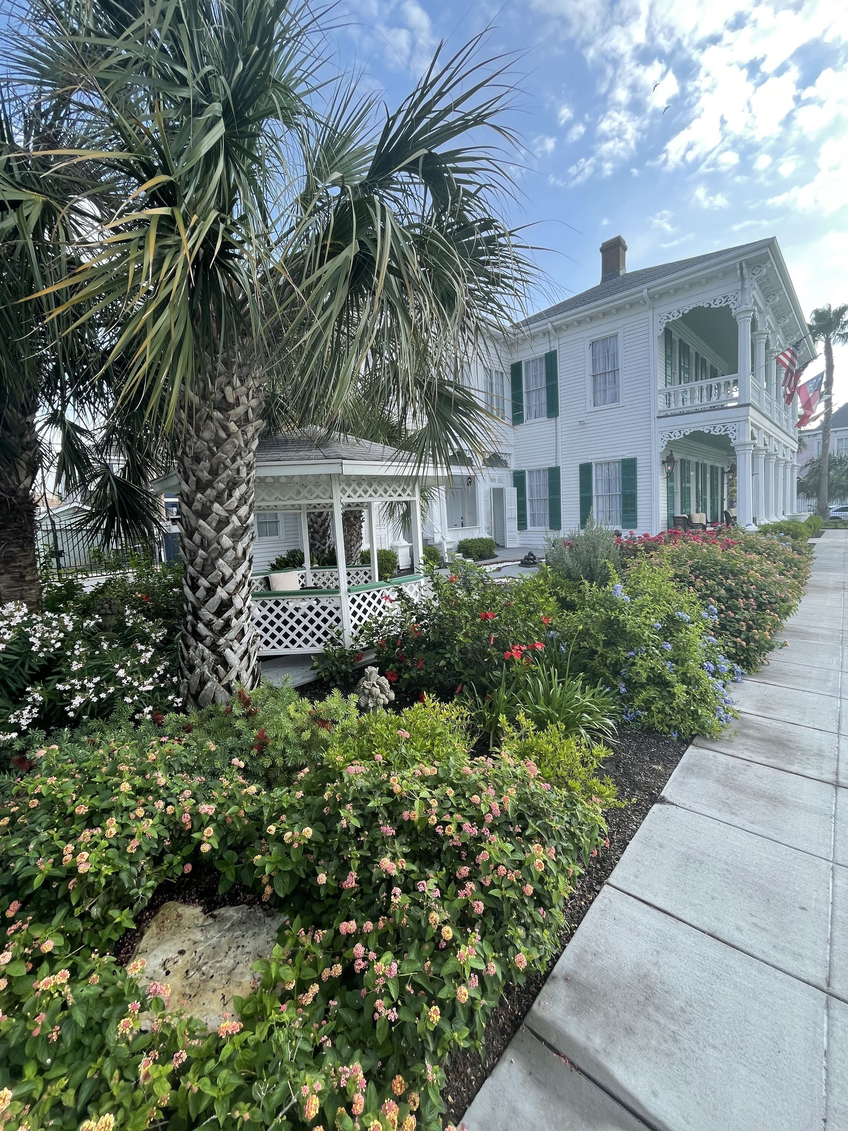 A white colonial-style building with a porch and a garden with various plants and flowers.