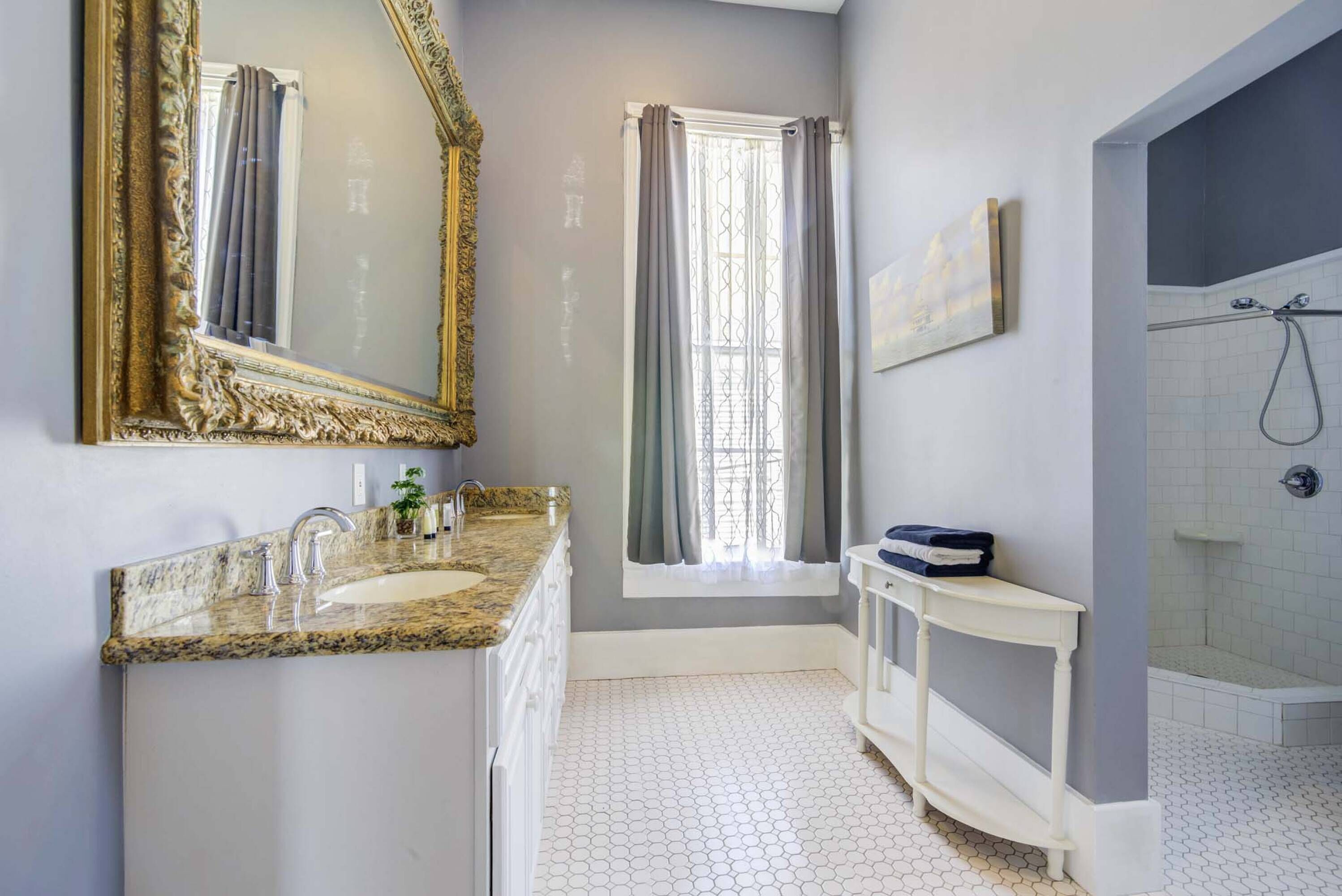 A bathroom with a granite countertop sink, a large ornate mirror, a window with curtains, a shower area, and a small white vanity.