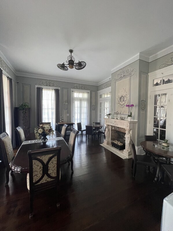 A spacious dining room with a high ceiling, ornate ceiling fan, and a fireplace with a mantel. - The George Manor (Galveston)