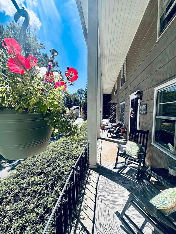 A covered porch with a hanging flower pot, outdoor seating, and a view of the house.