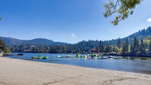 A beach with kayaks and rafts, a sandy shore, and a forested hillside in the background. - Lake & Mountain-View Home: Walk to Downtown! (Crestline)