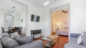 A modern living room with grey sofas, a glass coffee table, a TV mounted on the wall, and a dining area with a table and chairs. - Chic NOLA Villa: Pet-Friendly, Patio & Fire Pit (New Orleans)