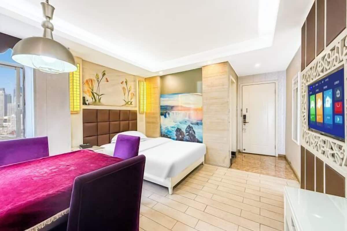 A hotel room with a bed, a dining table with chairs, a TV, and a city view.