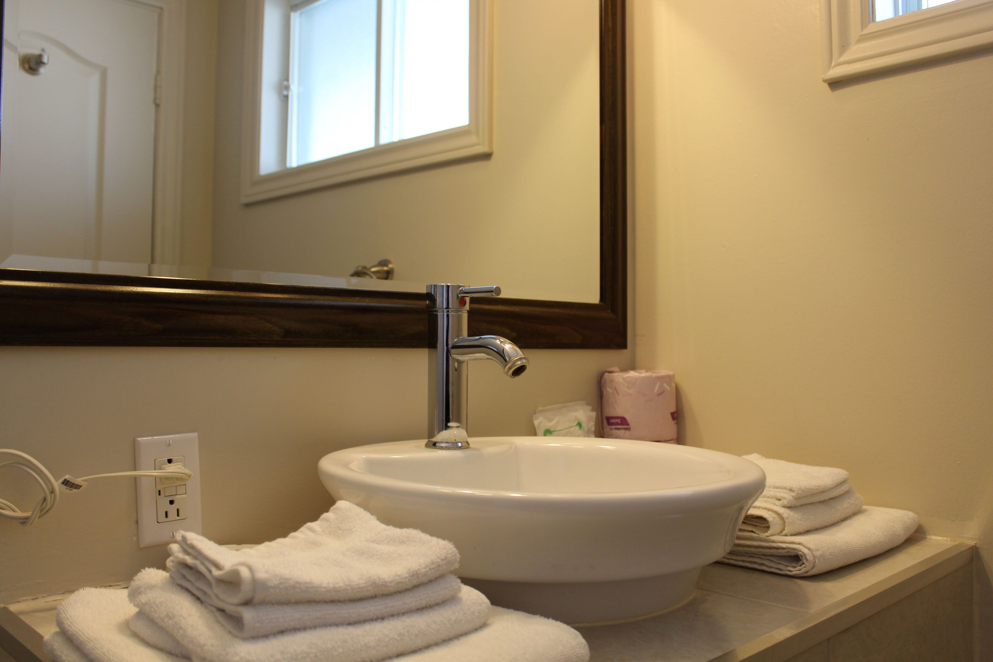 A bathroom with a white sink, a mirror, and a window above the mirror. There are towels stacked on the counter and a bottle of lotion.