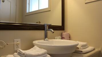 A bathroom with a white sink, a mirror, and a window above the mirror. There are towels stacked on the counter and a bottle of lotion.