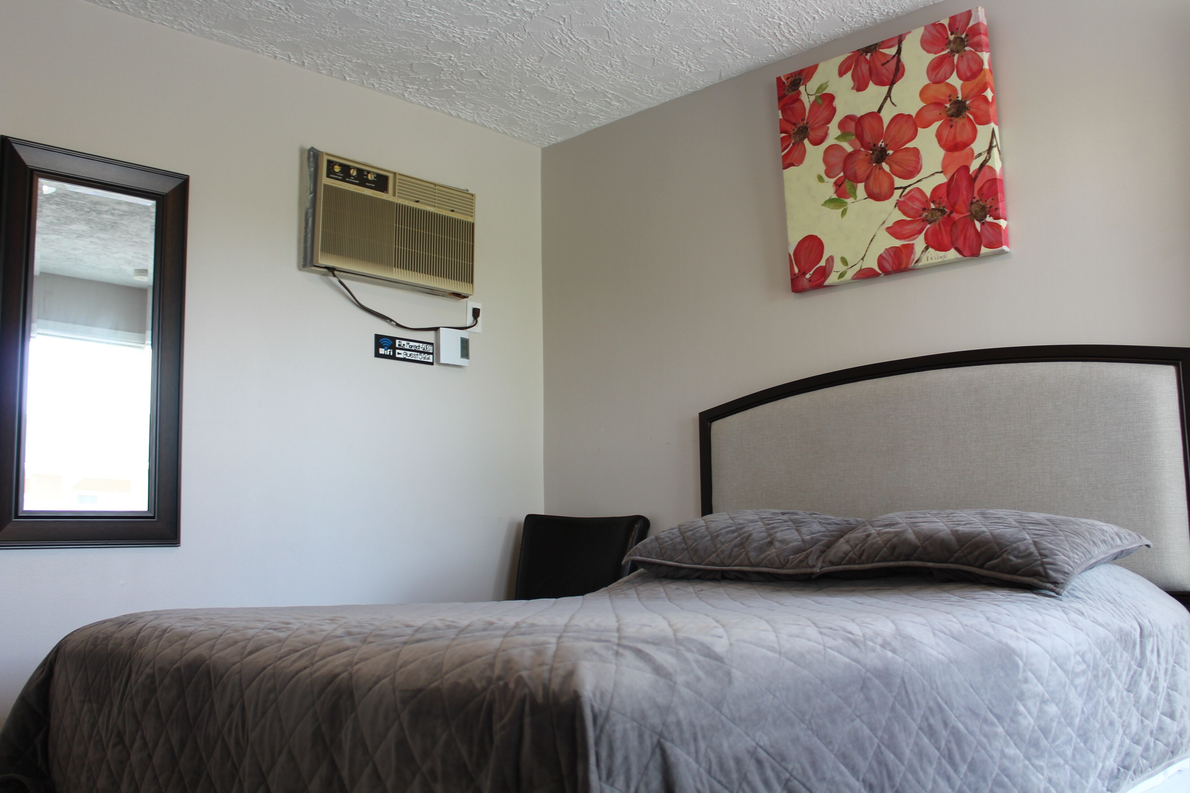 A bedroom with a bed, a window, an air conditioning unit, and a floral artwork on the wall.