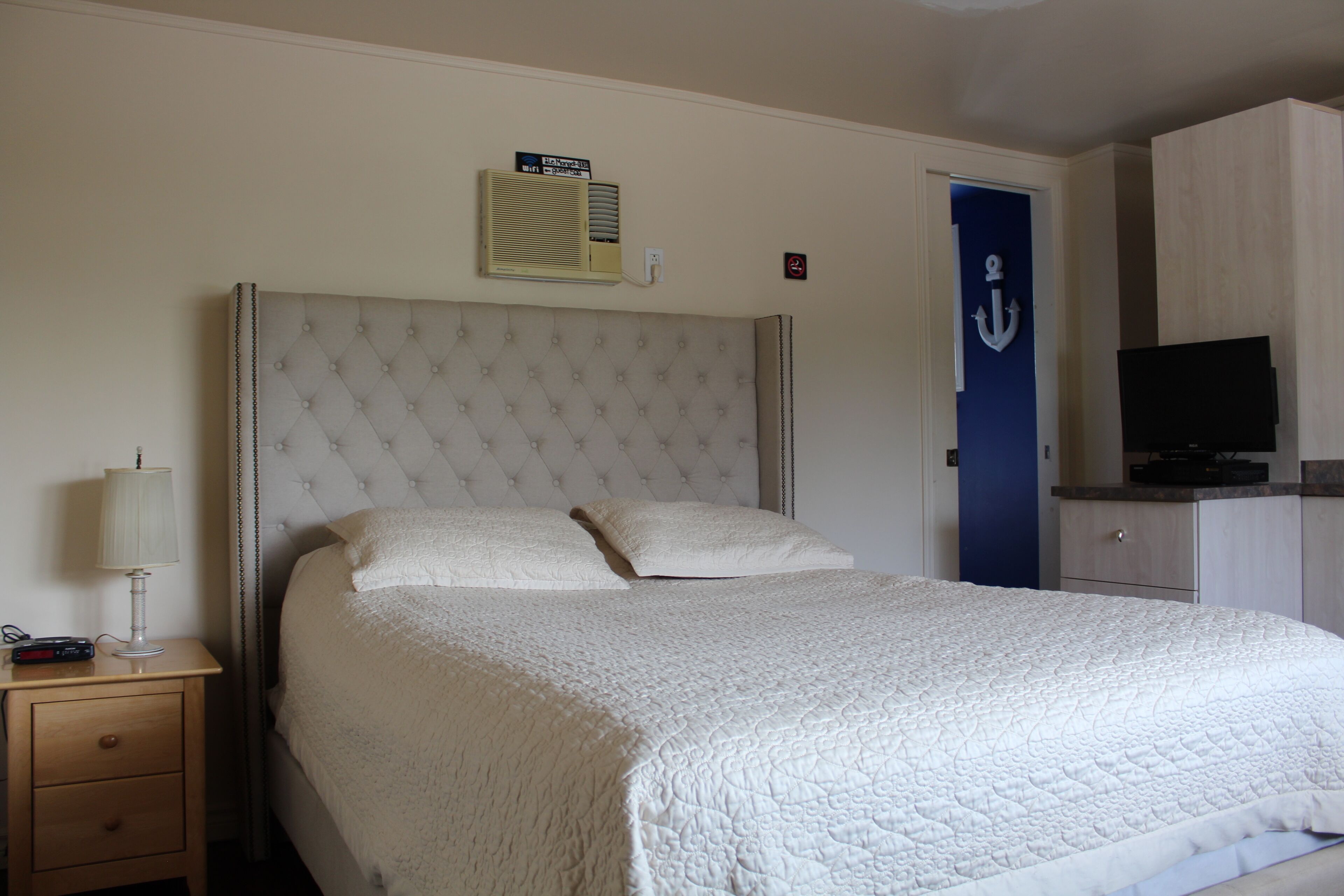 Ein Hotelzimmer mit einem großen Bett, einem Nachttisch mit Lampe, einem Fernseher und einer Tür mit Anker-Symbol.