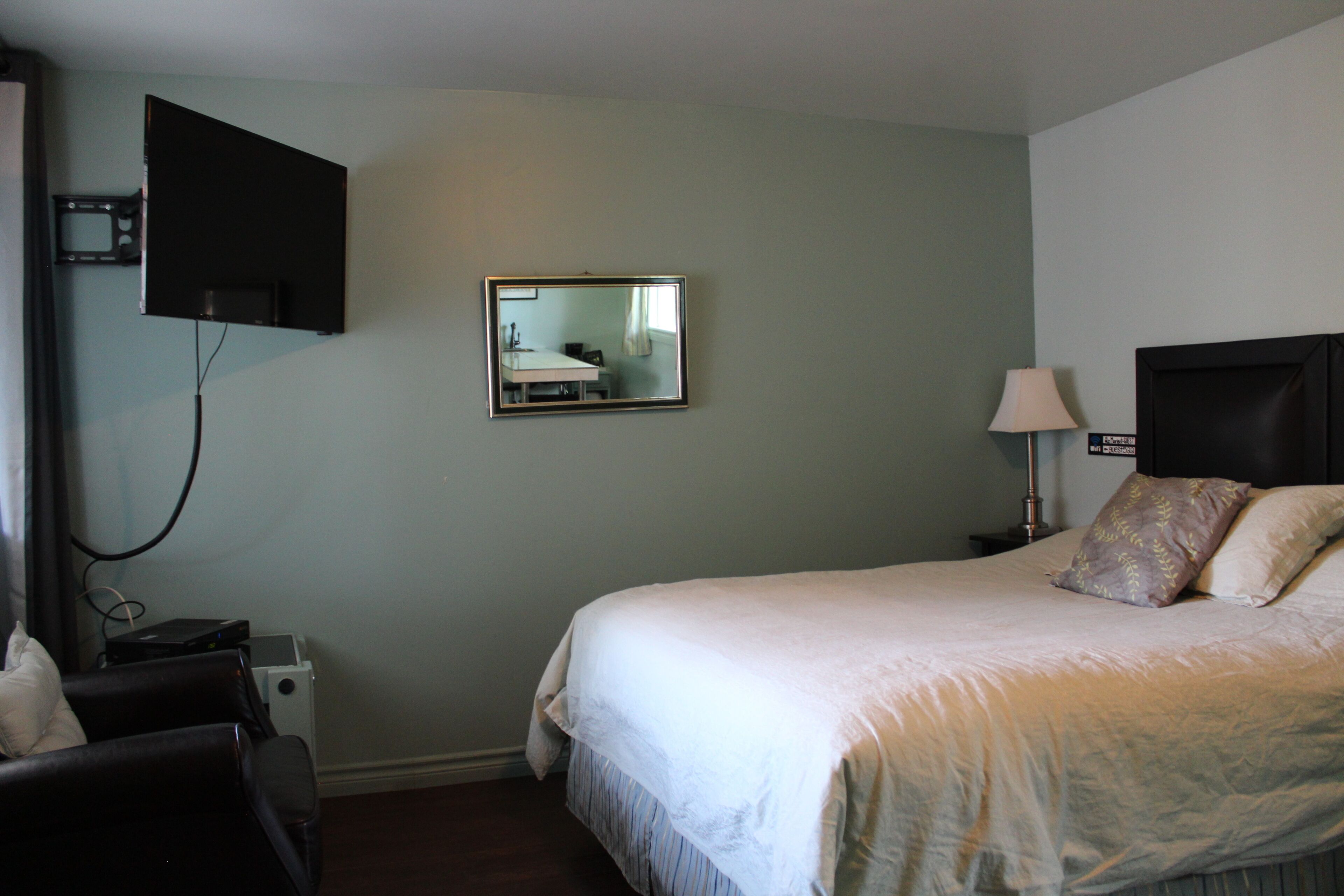 A hotel room with a bed, a TV mounted on the wall, a bedside table with a lamp, and a small desk with a chair.
