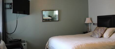 A hotel room with a bed, a TV mounted on the wall, a bedside table with a lamp, and a small desk with a chair.