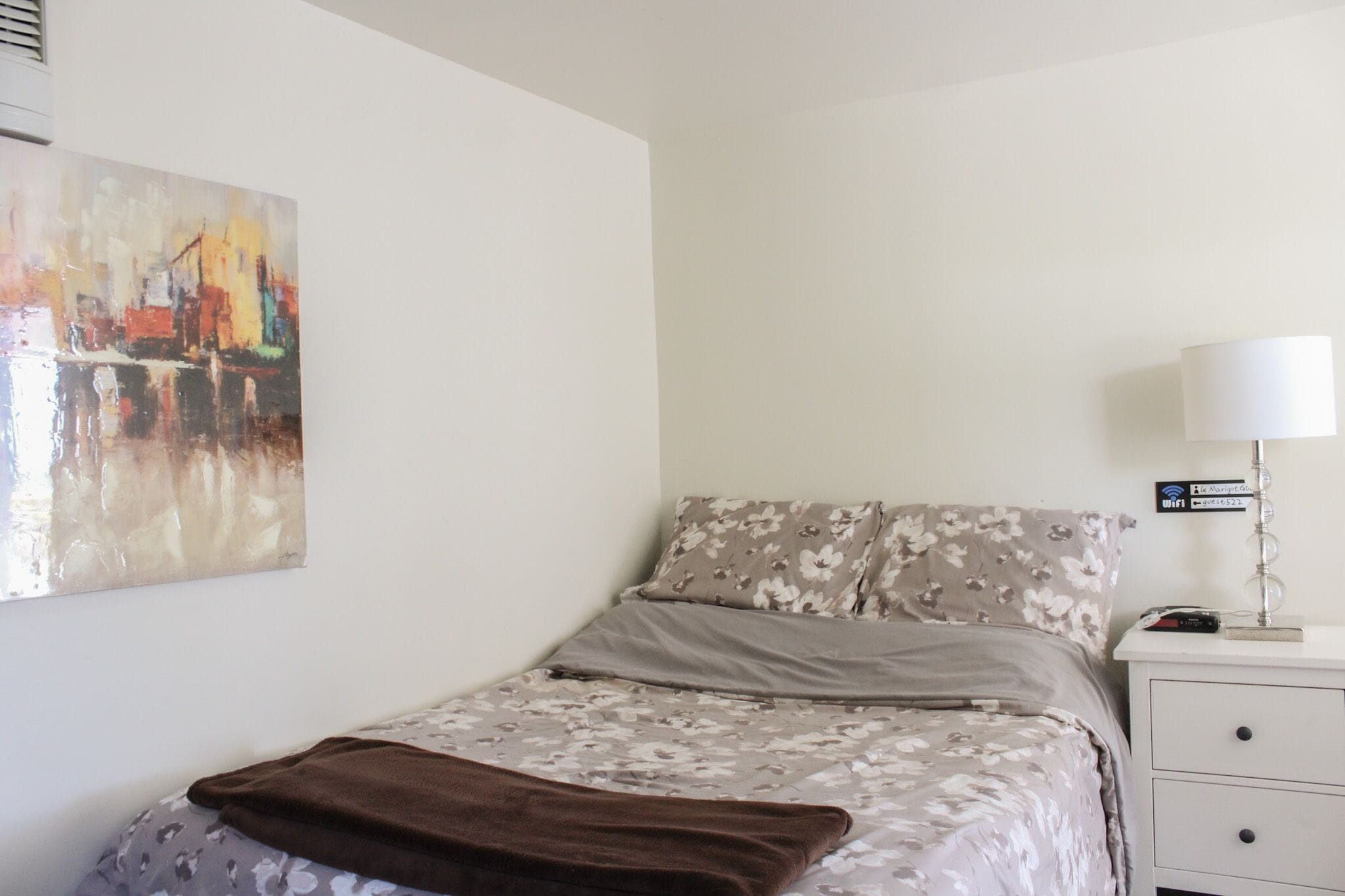 Un dormitorio con una cama, una mesita de noche, una lámpara, un cuadro e indicación de acceso a WiFi.