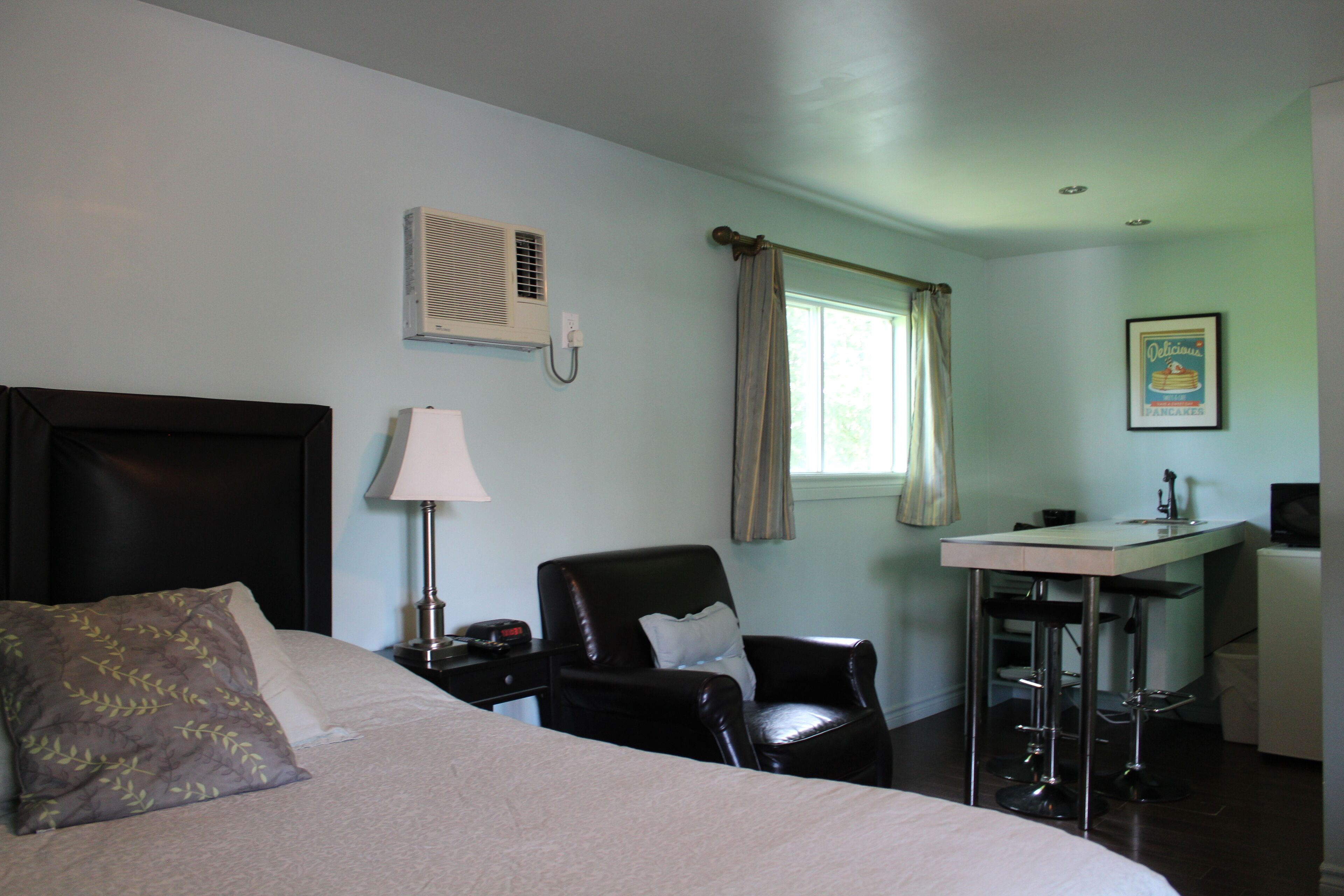 Habitación de hotel con cama, escritorio, silla, aire acondicionado, ventana con cortinas y un cuadro en la pared.