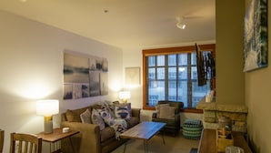 A cozy living room with a sofa, a coffee table, and a dining table. There is a window with blinds, a lamp, and a painting on the wall. - Grand Sierra Lodge: Walk to Village Gondola & Ski Back Trail (Mammoth Lakes)