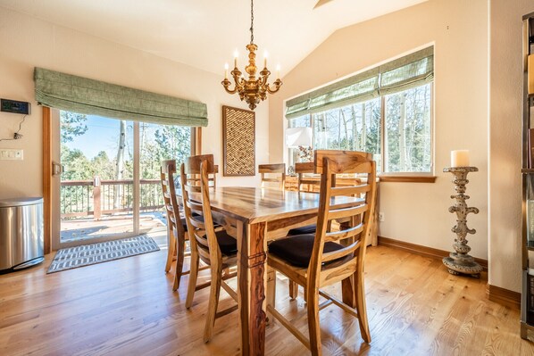 A dining area with a wooden table and chairs, a chandelier, and large windows with views of trees.