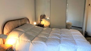 Una cama bien hecha con edredón blanco, una lámpara de noche y un espejo que refleja la habitación.