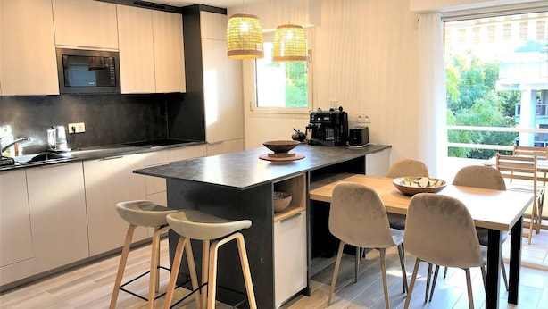 Una cocina moderna con una isla central, taburetes de bar, una mesa de comedor con sillas, una cafetera y un ventanal con vistas a zonas verdes.
